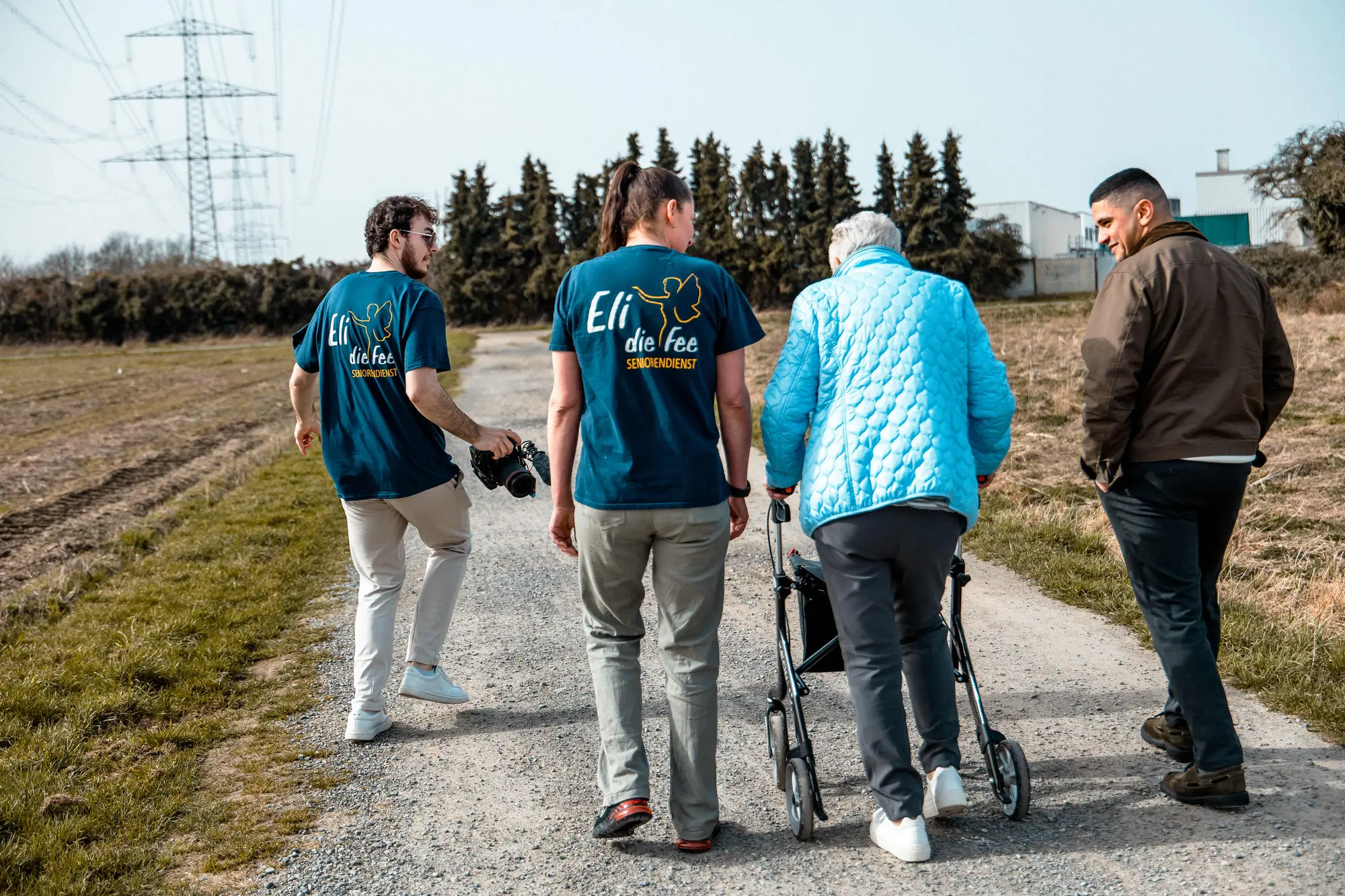 Betreuungskräfte von Eli die Fee gehen mit einer Seniorin am Rollator über einen Feldweg