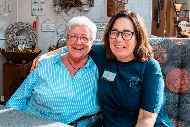 Betreuungskraft von Eli die Fee und eine Seniorin lachen gemeinsam auf dem Sofa