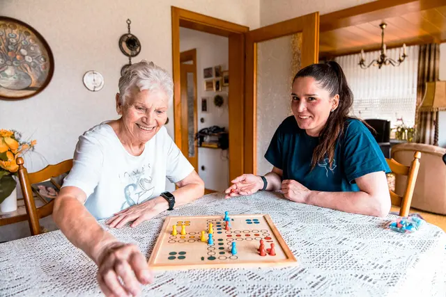 Seniorin und Betreuungskraft spielen am Wohnzimmertisch Mensch ärgere dich nicht