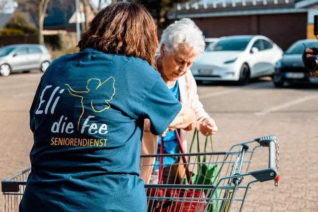 Betreuungskraft im Shirt von Eli die Fee hilft einer Seniorin beim Einkauf