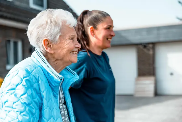 Seniorin in blauer Jacke lacht mit ihrer Betreuungskraft vor dem Haus
