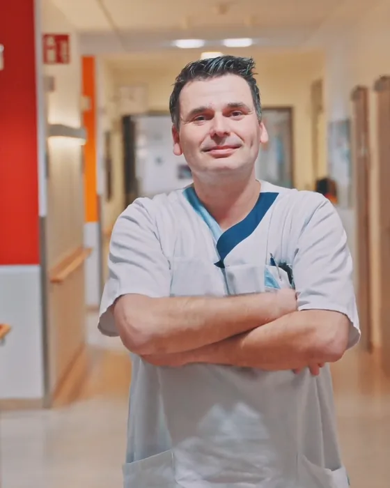 Emanuel in Dienstkleidung mit verschränkten Armen im Flur der Klinik